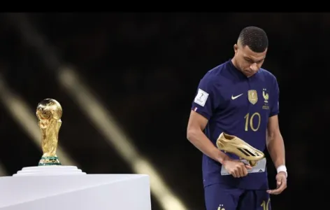 Kylian Mbappé com a medalha de prata no peito e o troféu da Chuteira de Ouro nas mãos, passando em frente à taça da Copa do Mundo