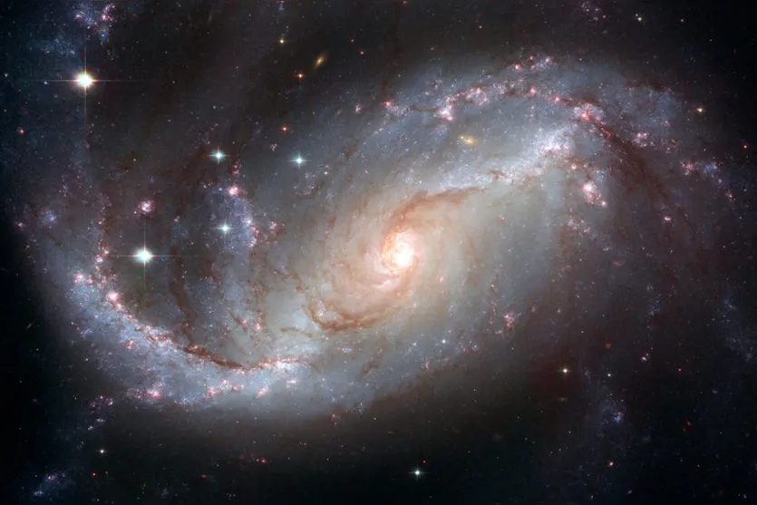   Galáxia NGC 1672 com grupos de estrelas e nuvens de gás hidrogênio brilhando em vermelho 